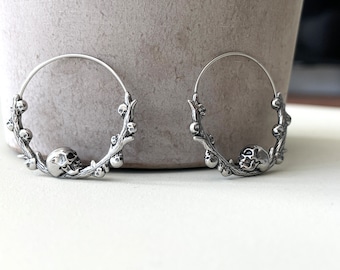 May include: Silver hoop earrings featuring a skull and branches with berries design.