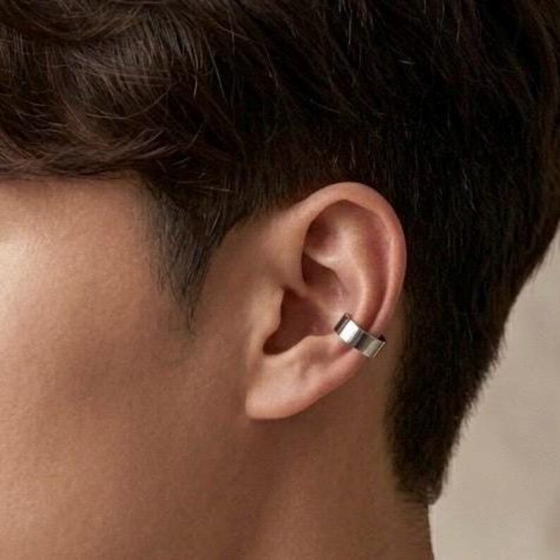 Pu&ograve; includere: Un ear cuff argentato &egrave; indossato sull'orecchio. L'ear cuff ha un design semplice e geometrico. La pelle della persona &egrave; marrone chiaro e i suoi capelli sono castano scuro. L'ear cuff &egrave; un accessorio moderno.