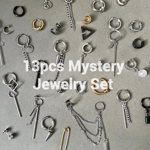 May include: A set of 13 silver and gold earrings with various designs, including chains, hoops, and studs. The earrings are arranged on a gray surface. 13pcs Mystery Jewelry Set