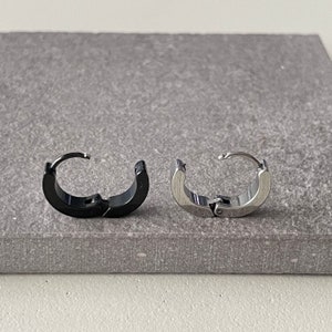 Pu&ograve; includere: Due orecchini a cerchio, uno nero e uno argentato, sono esposti su una superficie grigia. Gli orecchini hanno un design a cerniera. L'orecchino nero &egrave; lucido, mentre l'orecchino argentato ha una lucentezza metallica.