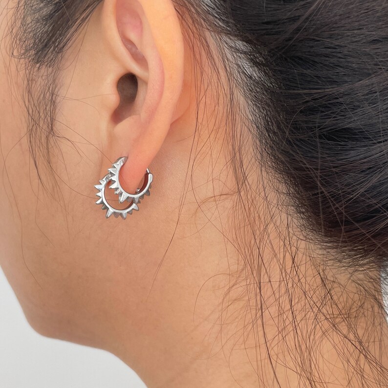 May include: A pair of silver hoop earrings with a spiky design. The earrings are worn on the ear, with the design facing outward. The earrings are a modern and edgy accessory.