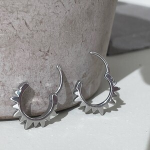 May include: A pair of silver-colored hoop earrings. Each earring features a series of triangular spikes along the outer edge. The earrings are designed to be hinged for easy wear and removal. The background is a neutral gray and white.