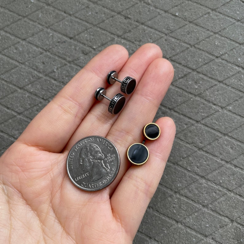 K&ouml;nnte beinhalten: Zwei Paar Ohrstecker. Ein Paar hat eine schwarze Mitte mit einem silbernen griechischen Schl&uuml;sseldesign am Rand. Das andere Paar hat eine schwarze Mitte mit einem goldfarbenen Rand. Eine US-Viertel-Dollar-M&uuml;nze ist ebenfalls im Bild.