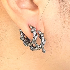 May include: A pair of silver-coloured hoop earrings. Each earring features a detailed snake design, with the snake's body coiled around the hoop. The snake's head is visible at the top and bottom of the hoop, creating a unique and edgy look.