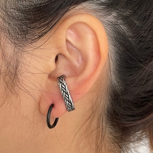 May include: A close-up of a person's ear with two earrings. The top earring is a silver, braided hoop. The bottom earring is a black, thin hoop.