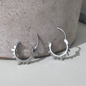 May include: A pair of silver-colored hoop earrings. Each earring features a series of triangular spikes along the outer edge, creating a sunburst design. The earrings are open, showing the clasp mechanism.