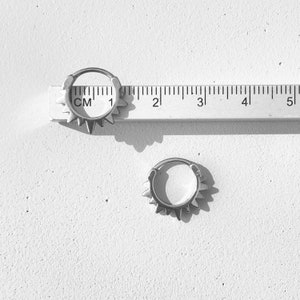 May include: Two silver-colored hoop earrings with a spiky design. The earrings are next to a ruler showing measurements in centimeters. The earrings are on a white surface.