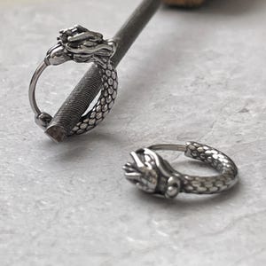 Könnte beinhalten: Zwei silberfarbene Creolen mit Drachenkopf-Details. Ein Ohrring befindet sich auf einer Metallfeile, der andere liegt auf einer hellgrauen Oberfläche. Der Drachenkopf befindet sich am oberen Ende des Reifens, mit einem strukturierten Körper.
