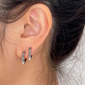 May include: A pair of small, silver hoop earrings. Each earring features a series of pointed, geometric designs along the outer edge. The earrings are worn on an ear, with the focus on the jewelry's design and texture.