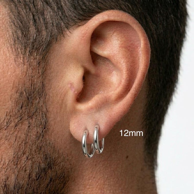 Puede incluir: Primer plano de una oreja con dos pendientes de aro plateados. Los pendientes tienen 1,2 cm de di&aacute;metro y son de metal brillante. La piel alrededor de la oreja es clara. El texto "12mm" es visible.