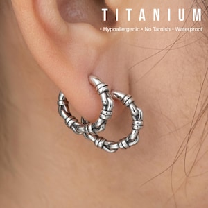 Puede incluir: Pendientes de aro plateados con un diseño similar al bambú. Los pendientes se llevan en una oreja, con la palabra "TITANIUM" y el texto "Hipoalergénico • No se empaña • Impermeable" visibles en el fondo.