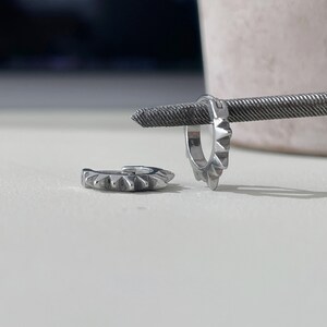 May include: Two silver hoop earrings with a spiked design. One earring is open, and the other is closed. The earrings are displayed on a white surface with a textured metal rod in the background.
