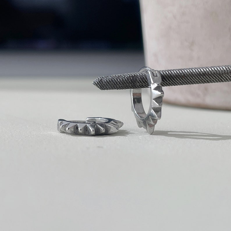 May include: Two silver hoop earrings with a spiked design. One earring is open, and the other is closed. The earrings are displayed on a white surface with a textured metal rod in the background.