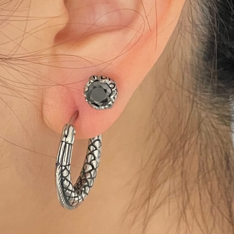 May include: A pair of silver earrings. One earring is a hoop with a textured snake design. The other earring is a stud with a black gemstone.