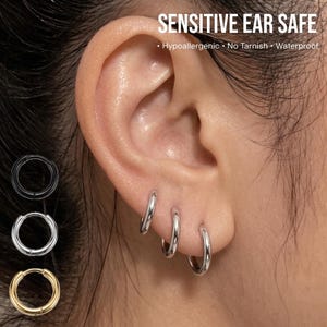 May include: Close-up of an ear with three small silver hoop earrings. The image also shows three additional hoop earrings in black, silver, and gold. Text at the top reads "SENSITIVE EAR SAFE" with additional text below.