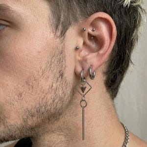 Puede incluir: Pendientes plateados con dise&ntilde;os geom&eacute;tricos. Uno presenta una forma de diamante con un colgante de cono y una barra vertical larga. El otro es un aro simple. Se ven m&uacute;ltiples perforaciones en la oreja.