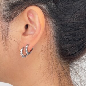 May include: A close-up of a person's ear wearing a pair of silver hoop earrings. The earrings have a spiky design around the outer edge. The person's hair is dark and pulled back.
