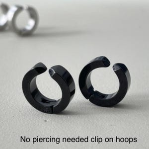 Pu&ograve; includere: Due orecchini a cerchio neri a clip sono mostrati su una superficie grigio chiaro. Gli orecchini sono circolari con un'apertura a cerniera. Il testo "No piercing needed clip on hoops" &egrave; nella parte inferiore dell'immagine.