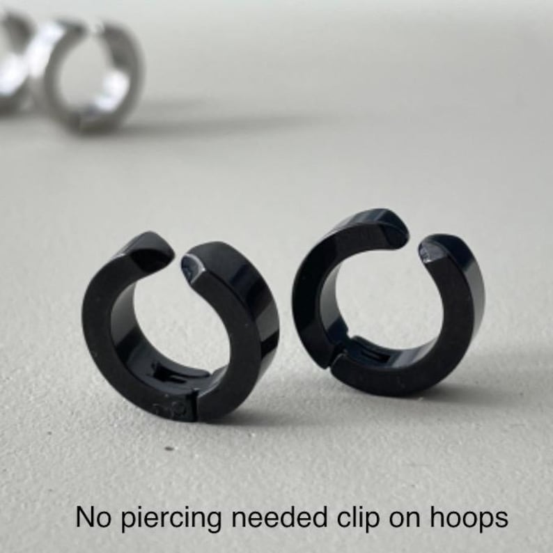 Pu&ograve; includere: Due orecchini a cerchio neri a clip sono mostrati su una superficie grigio chiaro. Gli orecchini sono circolari con un'apertura a cerniera. Il testo "No piercing needed clip on hoops" &egrave; nella parte inferiore dell'immagine.