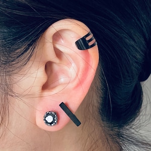 May include: A person's ear with three black ear cuffs. The ear cuff on the top of the ear is a simple bar design. The ear cuff in the middle of the ear is a small, round, black stone with a silver setting. The ear cuff on the bottom of the ear is a simple bar design.