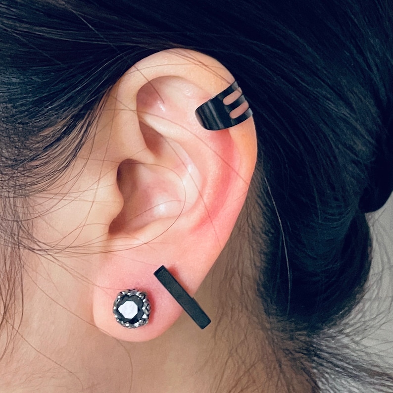 May include: A person's ear with three black ear cuffs. The ear cuff on the top of the ear is a simple bar design. The ear cuff in the middle of the ear is a small, round, black stone with a silver setting. The ear cuff on the bottom of the ear is a simple bar design.