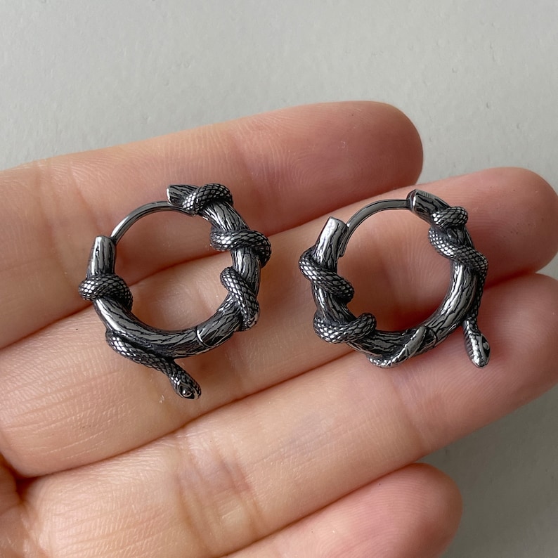 May include: A pair of silver-coloured hoop earrings. Each earring features a snake coiled around a textured, branch-like design. The snake's head and tail extend slightly from the hoop, creating a unique and edgy look. The earrings are designed for pierced ears.