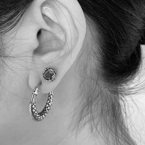 May include: A pair of silver earrings. One earring is a hoop with a textured snake design. The other earring is a stud with a black gemstone.