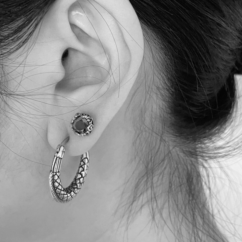 May include: A pair of silver earrings. One earring is a hoop with a textured snake design. The other earring is a stud with a black gemstone.
