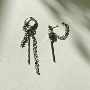 May include: A pair of silver earrings with multiple chain links and a small cube charm. One earring is a hoop with a dangling chain, the other is a stud with a dangling chain and cube charm.