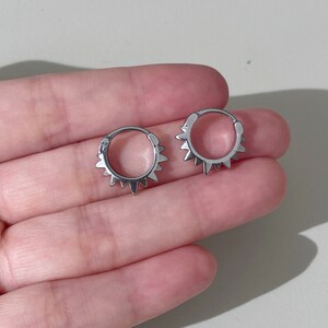 May include: Two silver-colored hoop earrings with a sunburst design. Each earring features a series of small, pointed spikes around the outer edge. The earrings are shown against a neutral background, held in a person's hand.