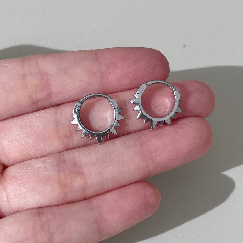 May include: Two silver-colored hoop earrings with a sunburst design. Each earring features a series of small, pointed spikes around the outer edge. The earrings are shown against a neutral background, held in a person's hand.