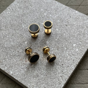 K&ouml;nnte beinhalten: Vier goldfarbene Ohrstecker mit schwarzen kreisf&ouml;rmigen Zentren. Zwei Paar Ohrringe sind sichtbar, jeweils in unterschiedlicher Gr&ouml;&szlig;e. Die Ohrringe haben ein griechisches Schl&uuml;sseldesign um die schwarze Mitte.