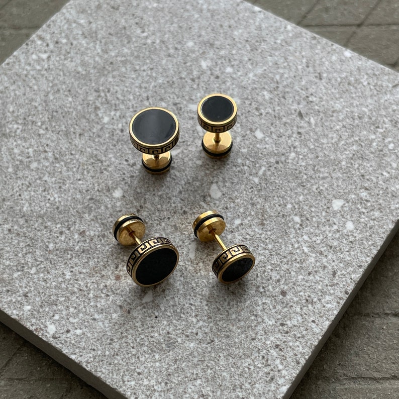K&ouml;nnte beinhalten: Vier goldfarbene Ohrstecker mit schwarzen kreisf&ouml;rmigen Zentren. Zwei Paar Ohrringe sind sichtbar, jeweils in unterschiedlicher Gr&ouml;&szlig;e. Die Ohrringe haben ein griechisches Schl&uuml;sseldesign um die schwarze Mitte.