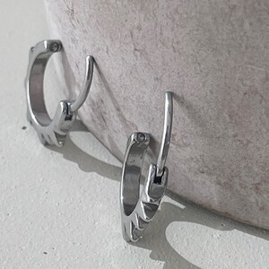 May include: Two silver-colored hoop earrings with a unique design. Each earring has a hinged closure and a decorative pattern along the outer edge. The earrings are displayed against a neutral background, highlighting their metallic sheen.