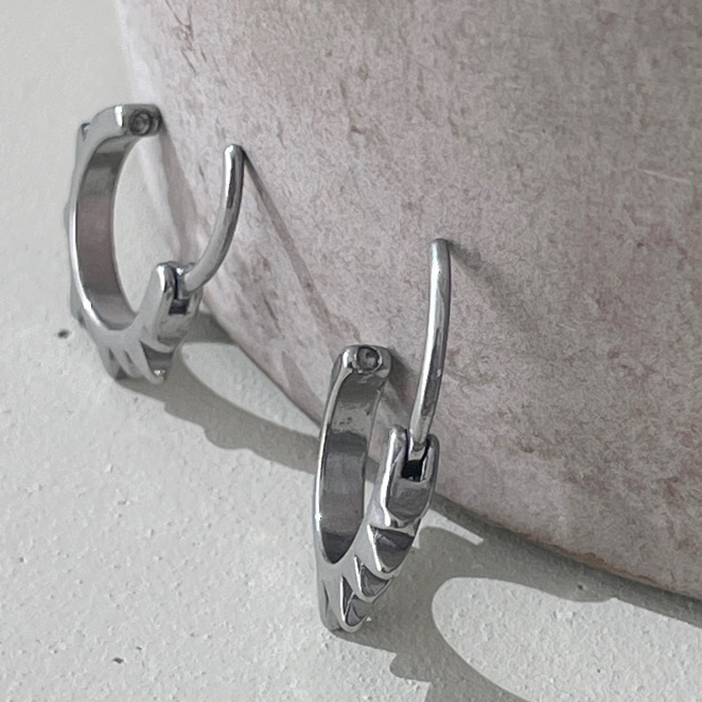 May include: Two silver-colored hoop earrings with a unique design. Each earring has a hinged closure and a decorative pattern along the outer edge. The earrings are displayed against a neutral background, highlighting their metallic sheen.