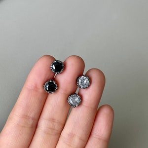 May include: Three silver stud earrings with a decorative border. Two earrings have black stones and one earring has a clear stone.