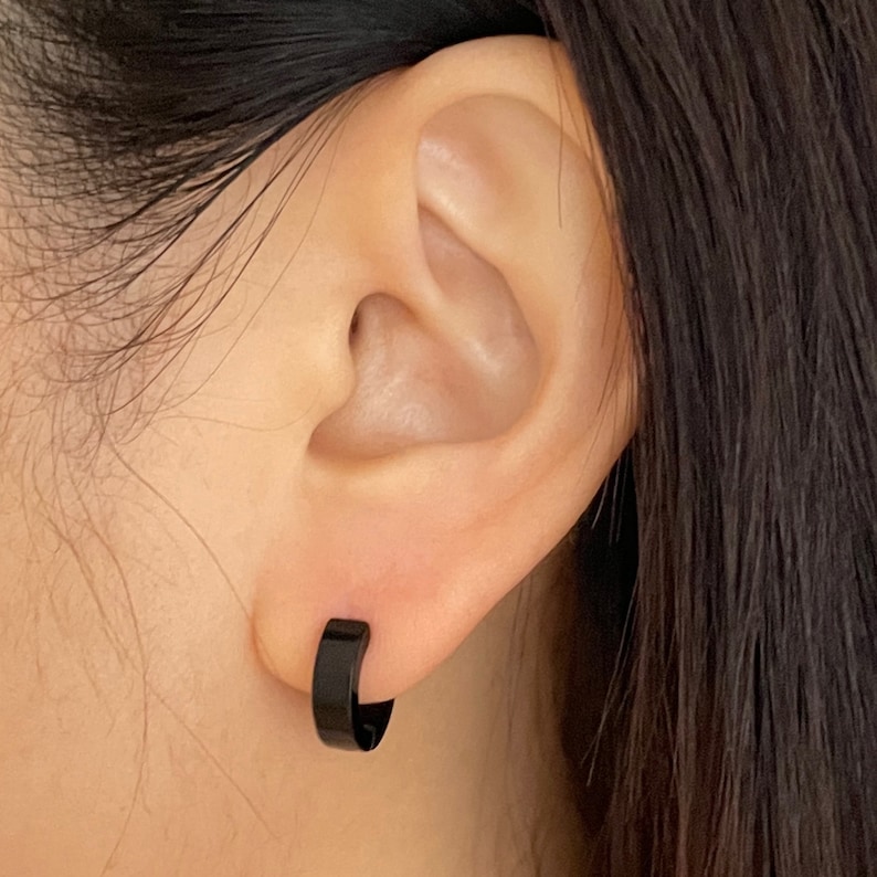 Pu&ograve; includere: Primo piano di un orecchio con un piccolo orecchino a cerchio nero. L'orecchino &egrave; una semplice fascia solida di metallo nero. La pelle intorno all'orecchio &egrave; liscia e chiara, con capelli scuri sullo sfondo.