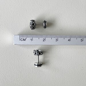 May include: Two silver earrings with a detailed design and black accents. The earrings are shown next to a ruler, measuring approximately 1 cm in length.