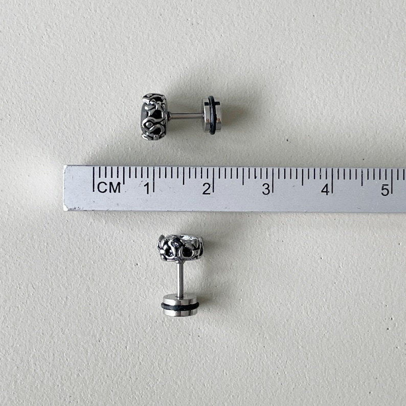 May include: Two silver earrings with a detailed design and black accents. The earrings are shown next to a ruler, measuring approximately 1 cm in length.