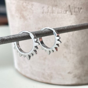 May include: A pair of small, silver hoop earrings with a sunburst design. Each earring features a series of triangular spikes around the outer edge. The earrings are displayed on a textured, metallic rod against a soft, neutral background.