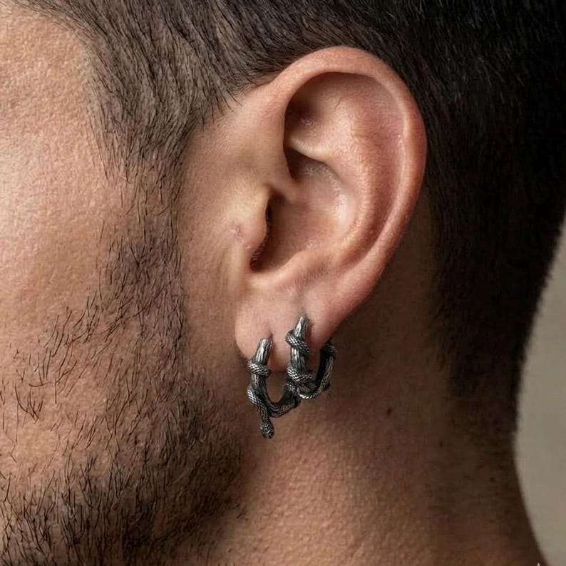 May include: Silver hoop earrings featuring a snake design. The earrings have a detailed, coiled snake pattern, giving a textured look. The jewellery is worn on the ear, highlighting the intricate design and metallic finish.