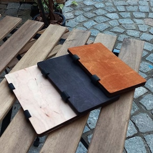 May include: Three wooden notebooks with black and brown covers. The notebooks are stacked on top of each other on a wooden surface.
