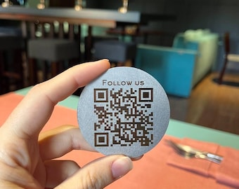 May include: A round, silver-coloured coaster with a black QR code and the text "Follow us" printed on it.