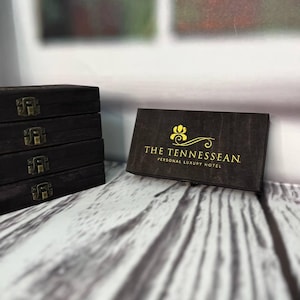 Puede incluir: Una caja de madera marrón oscuro con un logotipo dorado que dice "THE TENNESSEEAN. PERSONAL LUXURY HOTEL".
