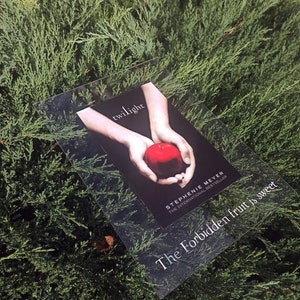 May include: A book cover with the title "Twilight" by Stephenie Meyer, the international bestseller. The cover features a hand holding a red apple with the text "The Forbidden fruit is sweet" below.