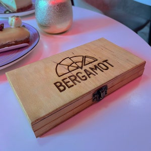 May include: A wooden box with a hinged lid and a brass latch. The box is engraved with the word "BERGAMOT" and a stylised image of a citrus fruit.