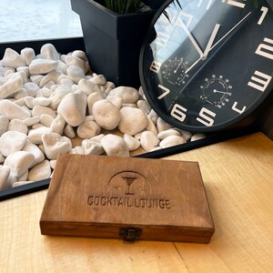 May include: A wooden box with a brown stain and a brass latch. The box has a logo that says "Cocktail Lounge" and a stylized image of a cocktail glass.