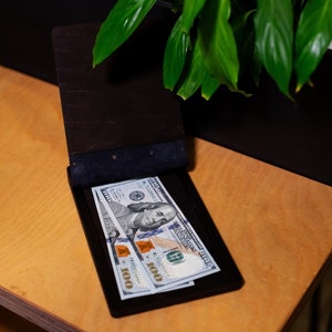 Op de afbeelding: Twee biljetten van $ 100 liggen in een zwarte leren portemonnee met een blauwe suède voering. De portemonnee is open en de biljetten zijn zichtbaar. De portemonnee ligt op een lichtbruin houten oppervlak.