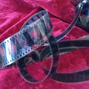 May include: A coiled strip of 35mm photographic film featuring translucent dark frames with visible sprocket holes along the edges. The film strip is transparent and reflects light, showing faint imagery within the individual rectangular frames.