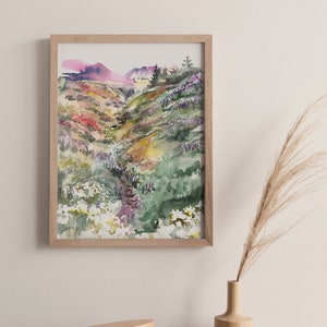 May include: A framed watercolor painting of a green, yellow, and purple landscape with a winding path. The painting is hanging on a white wall above a wooden dresser with a small wooden box and a vase with dried grass.